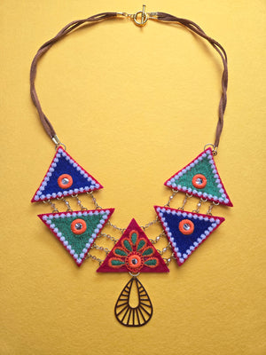 Embroidered Harmony Crystal Triangle Necklace Five two inch Colorful mix and match embroidered triangle shapes within a 26 inch long statement necklace, of green, blue, and red, with hints of white and orange, each with a mirrored center, and arranged in a pattern that is connected by chains with glass crystals. Closure is a toggle clasp on loosely twisted tan sueded leather. A brushed brass tear drop cut-out pendant is connected under the center triangle. 