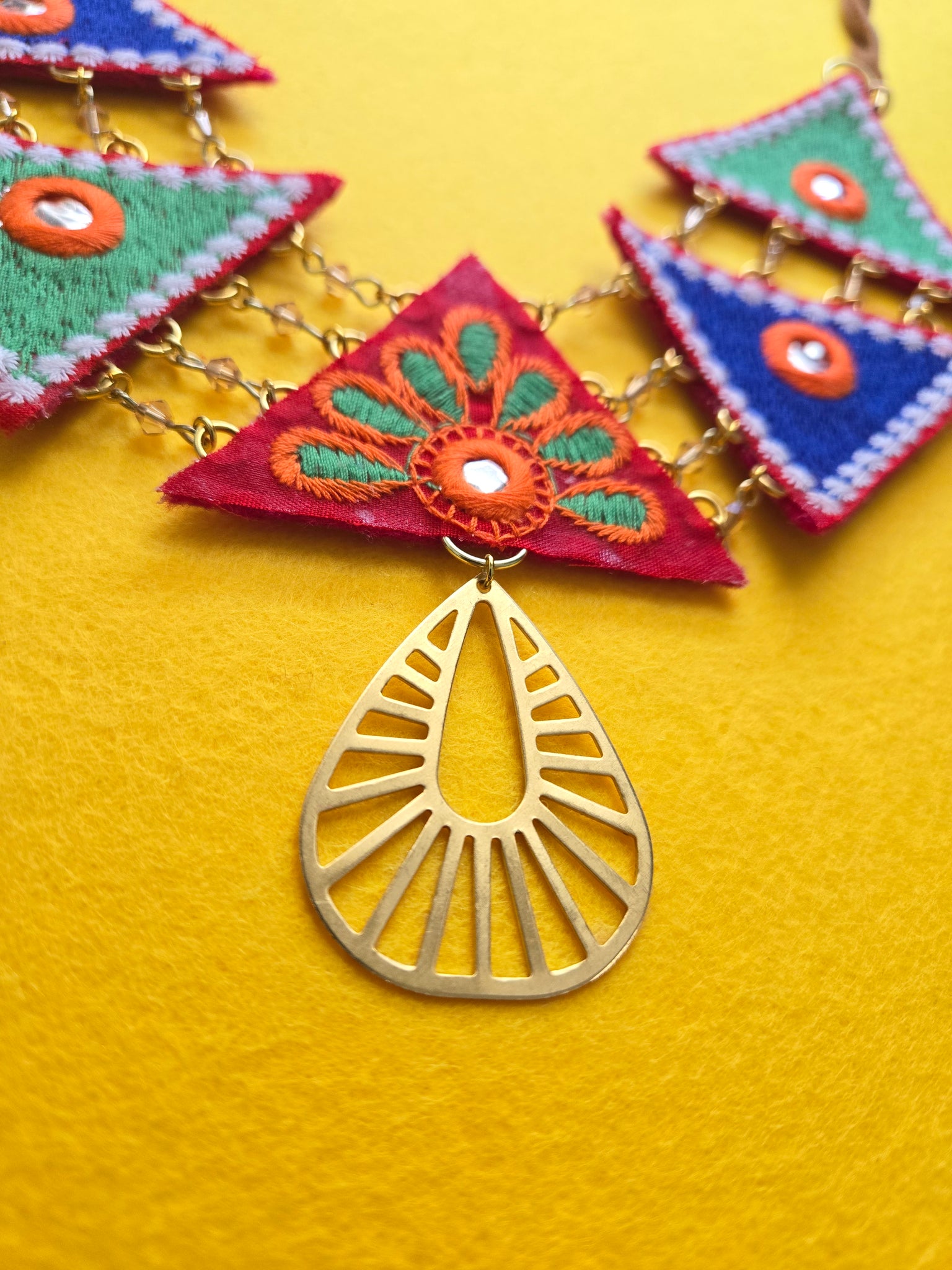 Close up of Necklace with five two inch colorful embroidered triangles suspended and separated by gold chain. Each triangle is a different color of blue, green, or red. Necklace is also hung on sueded leather, and has a cut-out brass pendent at the bottom of the triangle pattern.