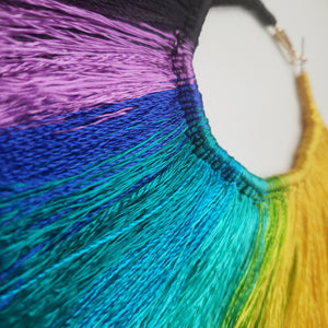 This image shows a close up of how the colors of the earrings look. Fan shaped Multi-Colored Tassel Hoop earrings that are yellow, lime-green, blue, purple, and black. Made with satin thread